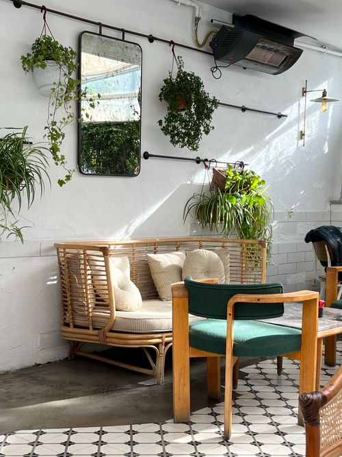 Free Sunlit café corner with rattan sofa and houseplants for a cozy and modern vibe. Stock Photo