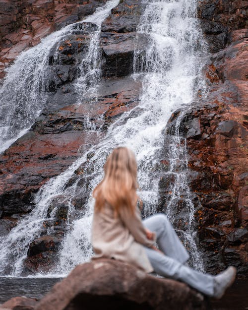 Free A Woman Sitting on a Rock Near Waterfall Stock Photo