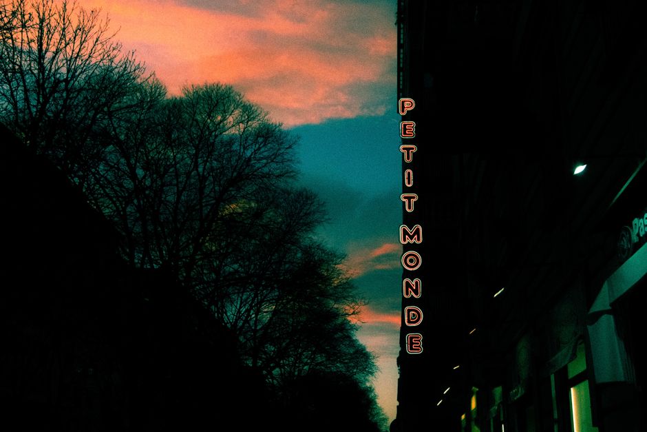 Urban skyline at dusk featuring 'Petit Monde' neon sign against a vibrant sunset sky.