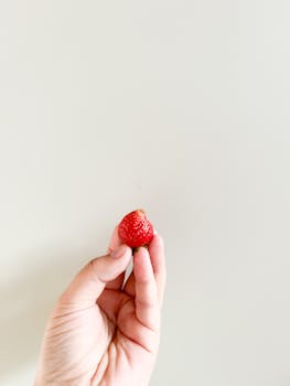 Soft focus image of a hand holding a fresh strawberry against a minimalist background.