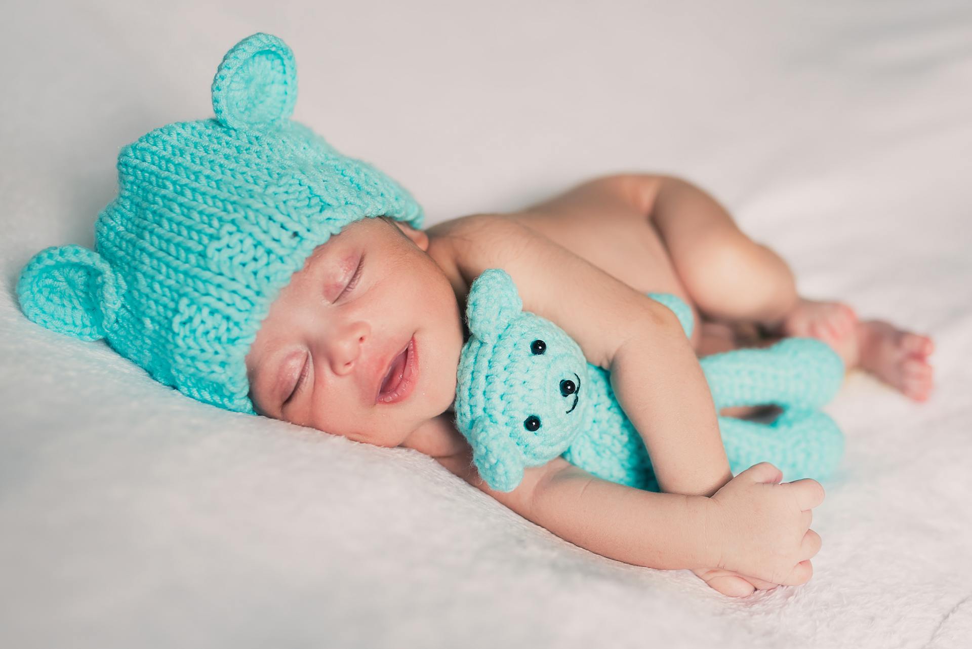 Photo of a Newborn Wearing a Cap and Keeping a Teddy