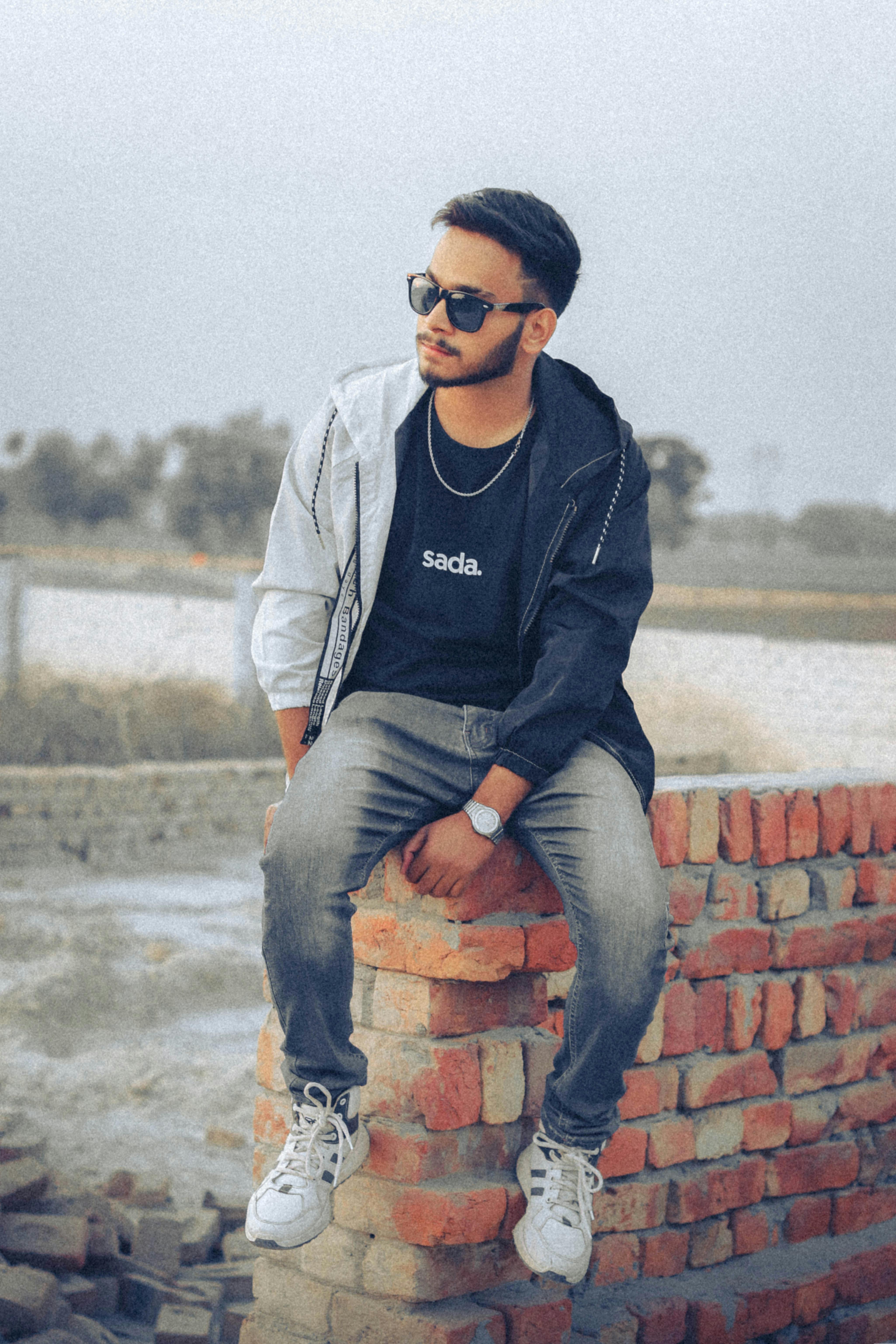 Fashionable young man sitting stylishly on a brick wall with sunglasses outdoors.