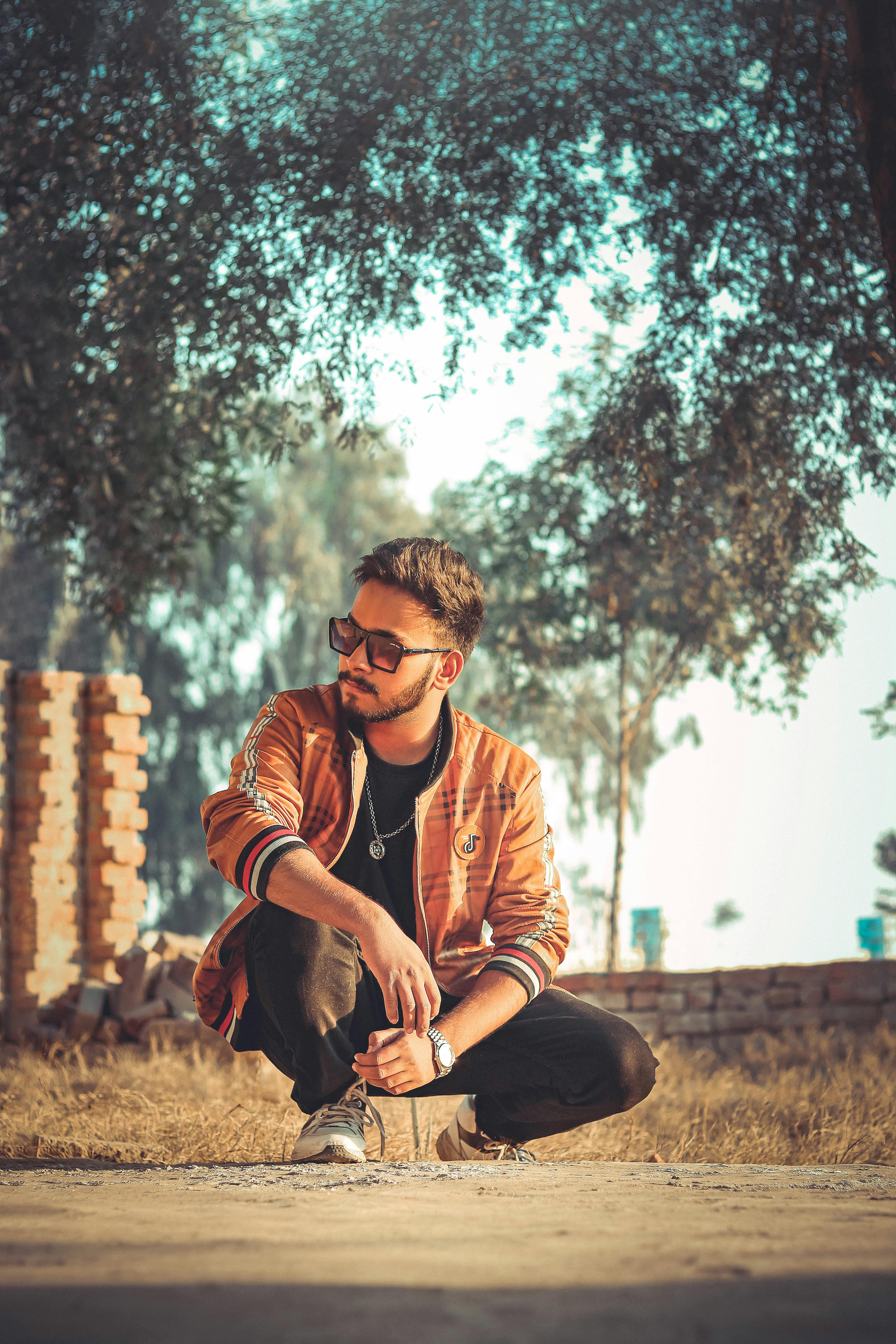 A fashionable man in sunglasses crouches stylishly outdoors with sunlight filtering through trees.