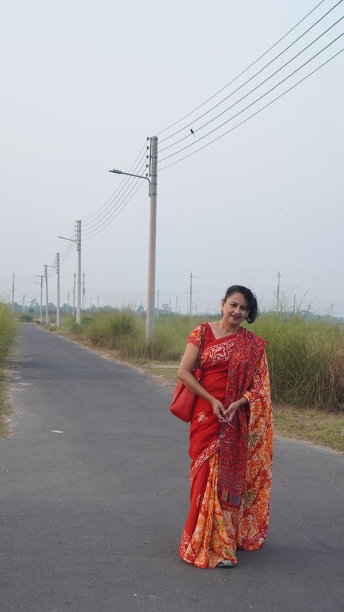 Free Woman in a vibrant red sari stands on a rural road lined with electric posts, capturing cultural elegance. Stock Photo