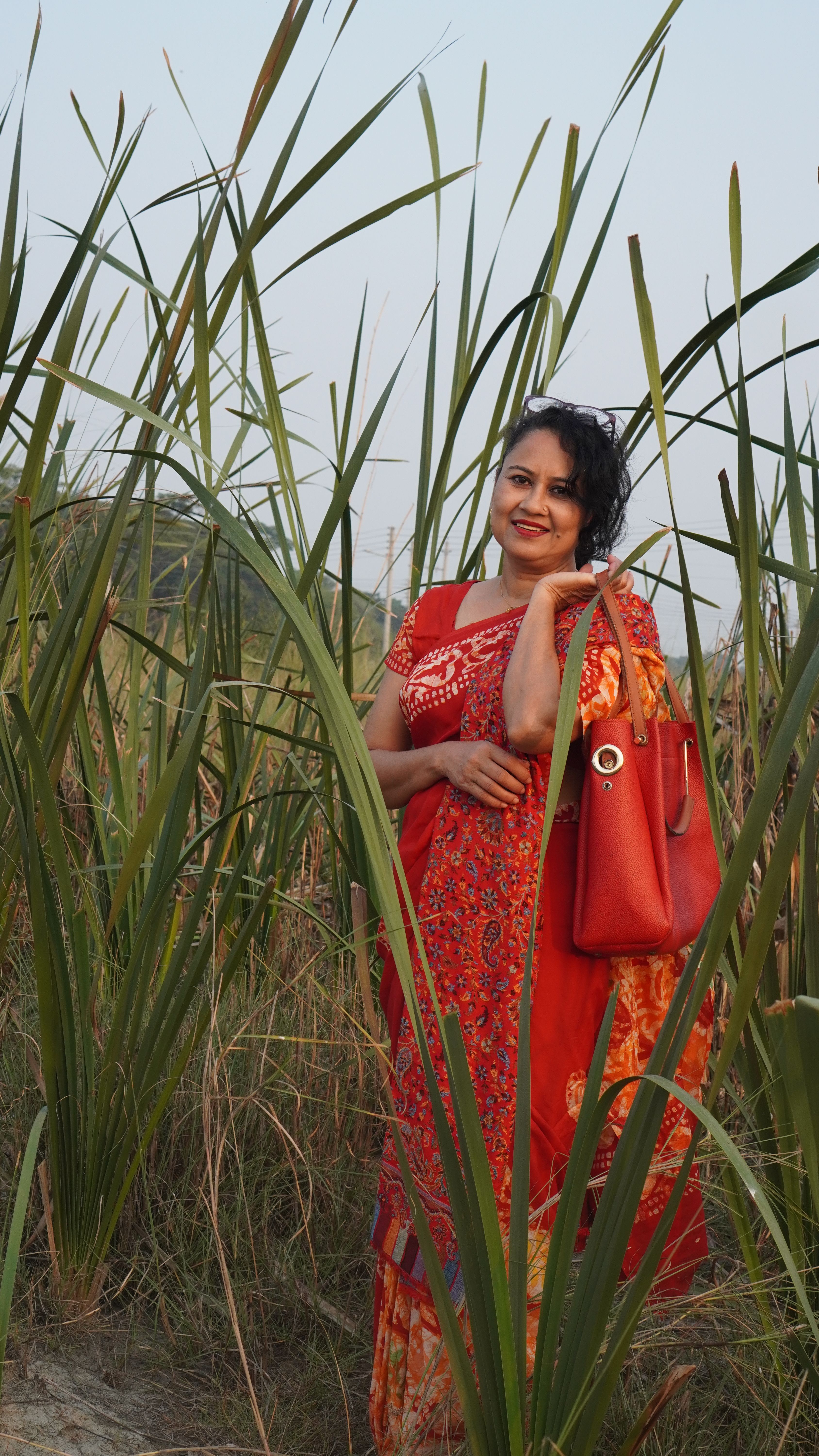 Woman in a vibrant red traditional dress posing amidst tall green plants outdoors.