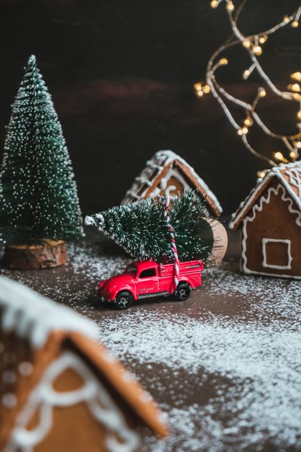 Miniature red truck carries Christmas tree in gingerbread village scene.