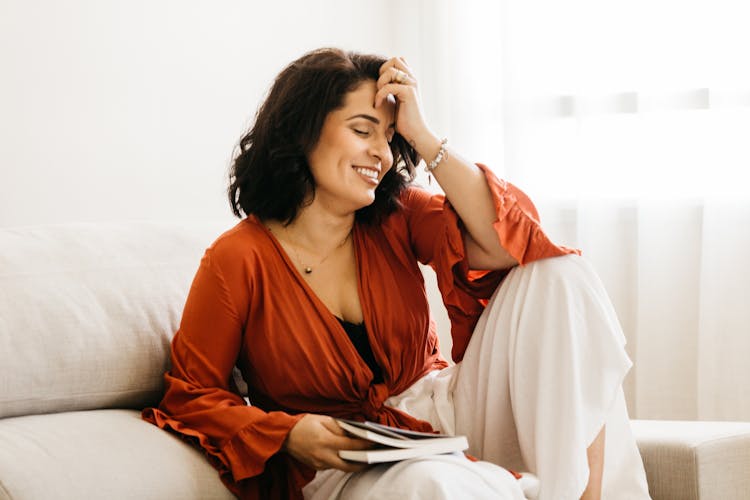 Photo Of A Smiling Woman With Hand Raised, Sitting On A Sofa And Keeping A Book