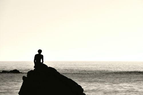 Free A person sits silhouetted on rocks by the ocean at sunset near California's coastline. Stock Photo