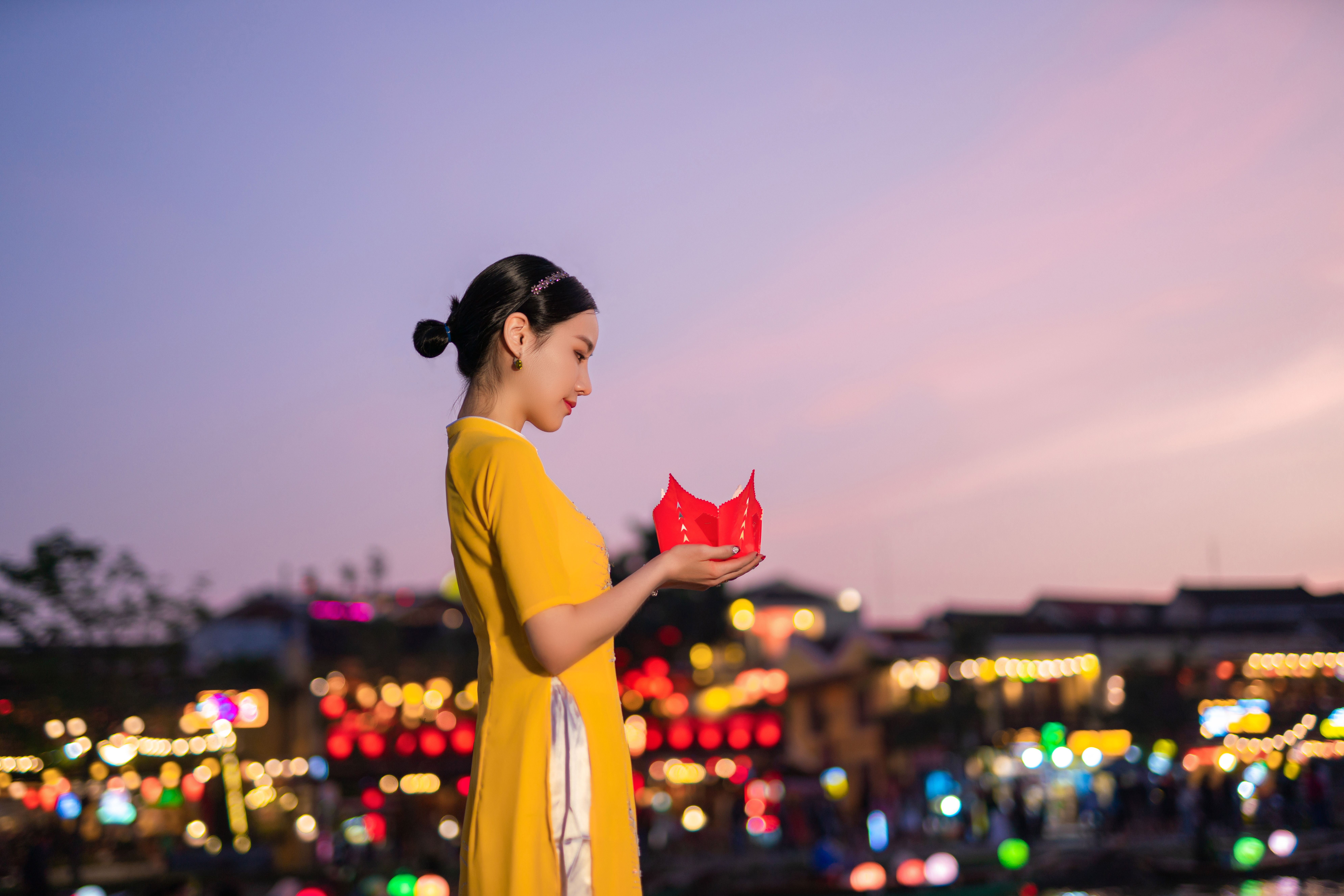 An Asian woman in a yellow dress holds a lantern amidst a vibrant, festive setting at twilight.