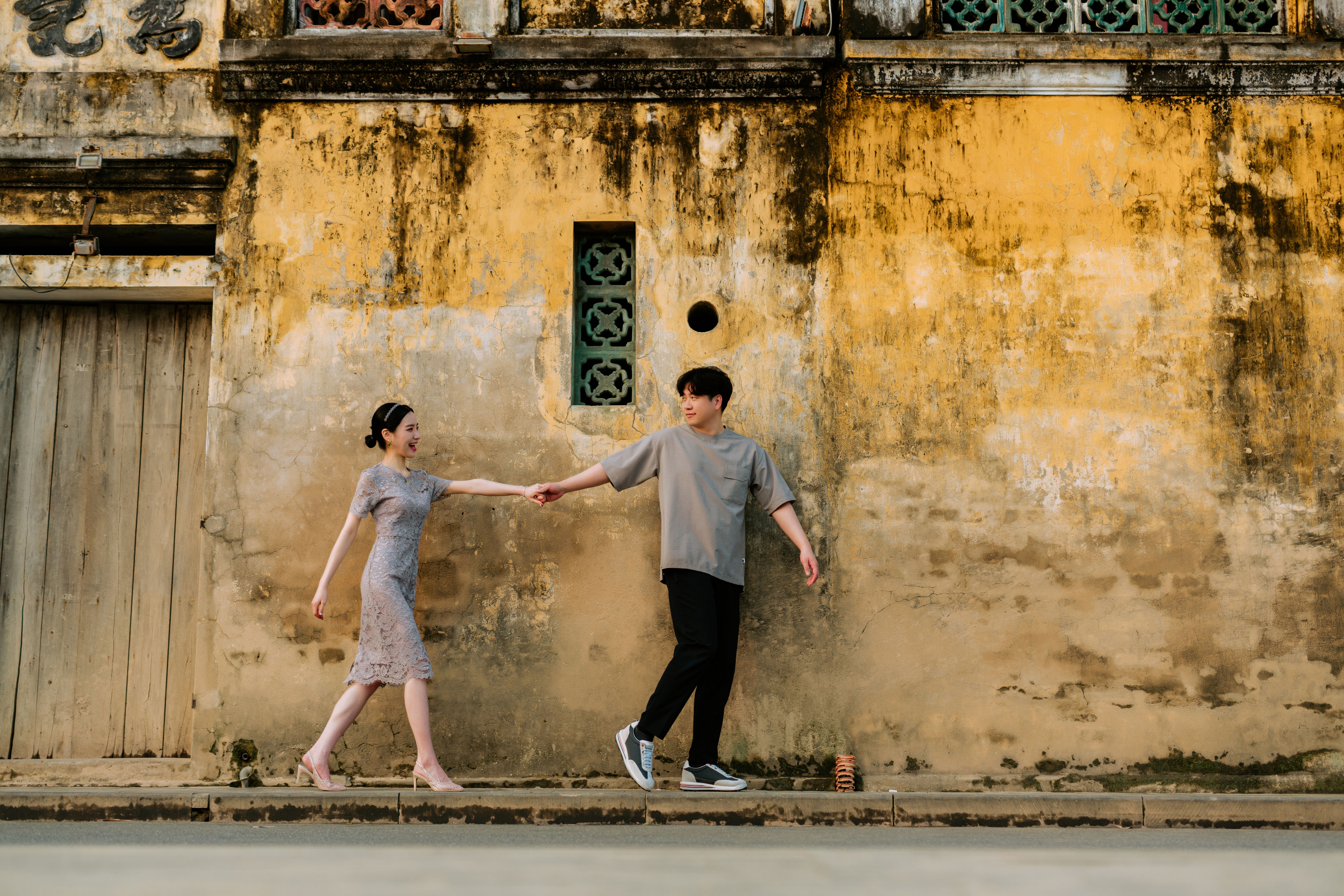 A charming couple walking hand in hand against a rustic wall, showcasing love and affection.