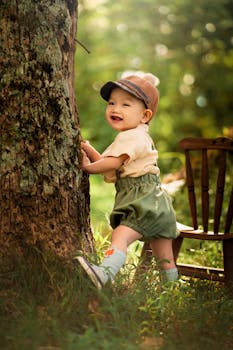 Adorable baby girl in a charming outfit standing by a tree outdoors.