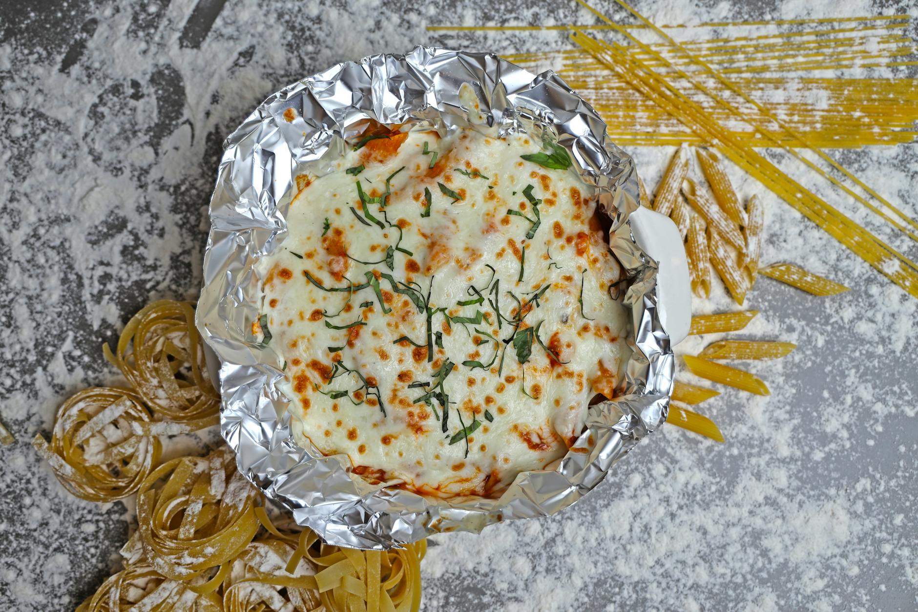 Delicious baked pasta topped with cheese and basil, surrounded by uncooked pasta and flour.