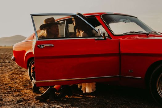 Two people in cowboy hats next to a vintage red car at sunset on a barren landscape in Turkey.