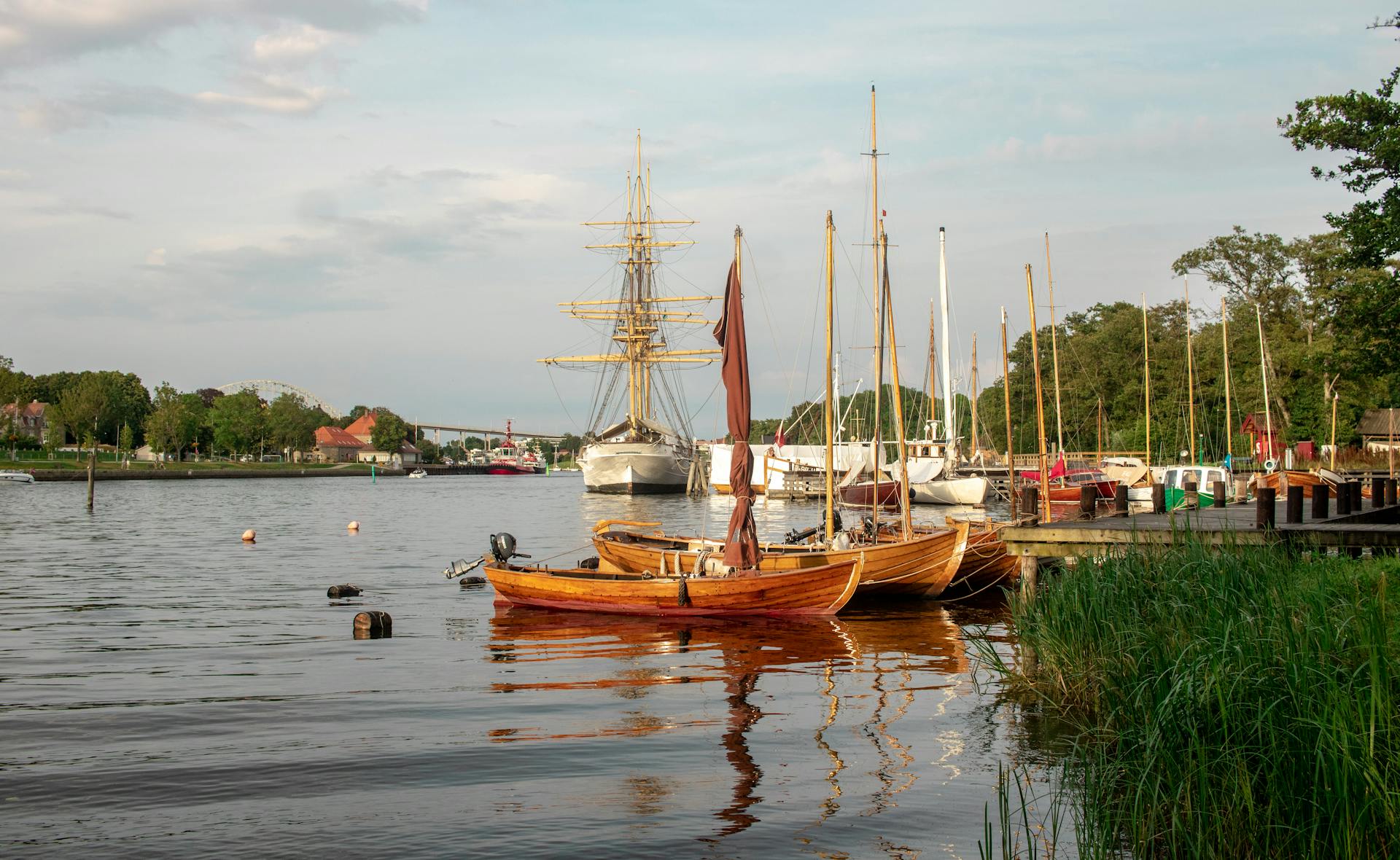 A peaceful harbor scene with moored wooden sailboats reflecting on calm water under a soft, cloudy sky.