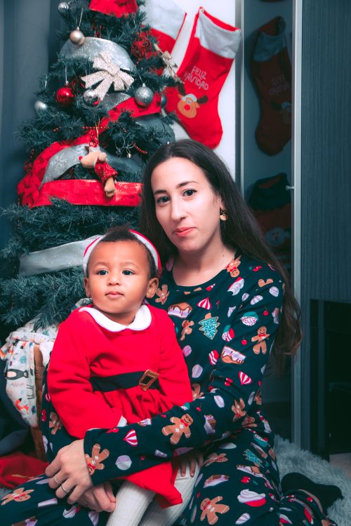 Free A mother and her baby in festive attire sitting by a Christmas tree, enjoying the holiday spirit indoors. Stock Photo