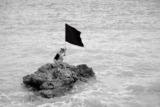 Woman kneeling on rock holding black flag against ocean backdrop, in a monochrome photo.