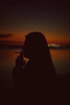 Artistic silhouette of a person smoking against a vibrant twilight background over water.