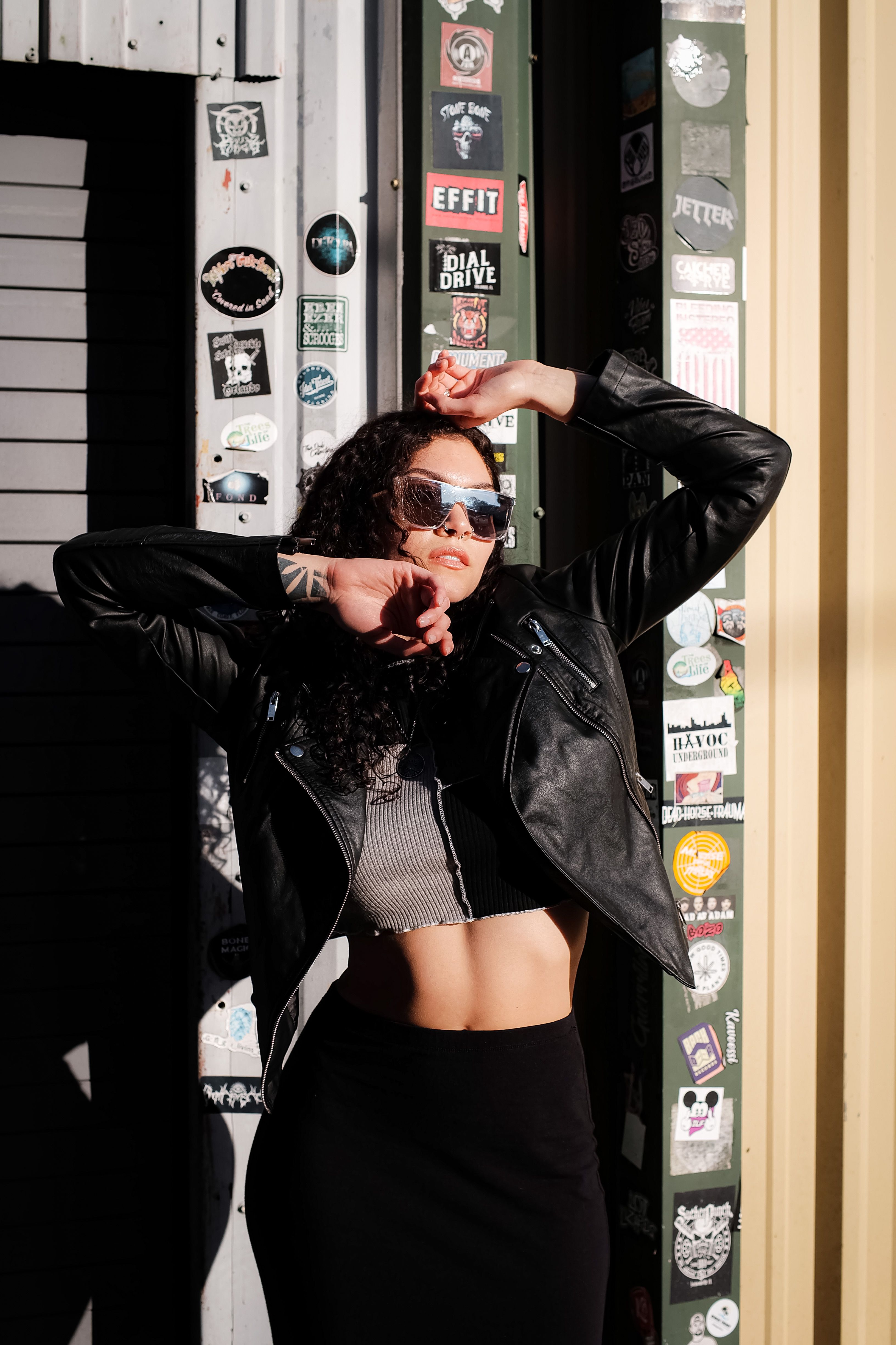 Fashionable woman in sunglasses posing against sticker-covered wall in sunlight.