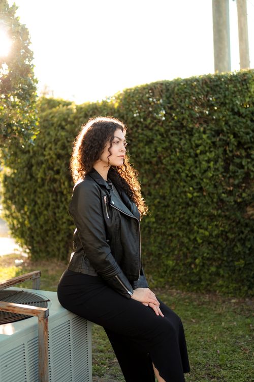 Free A woman in a leather jacket sitting outdoors on a sunny day, surrounded by greenery. Stock Photo