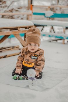 Smiling baby bundled up warmly, playing in the snow with a small snowman during winter.