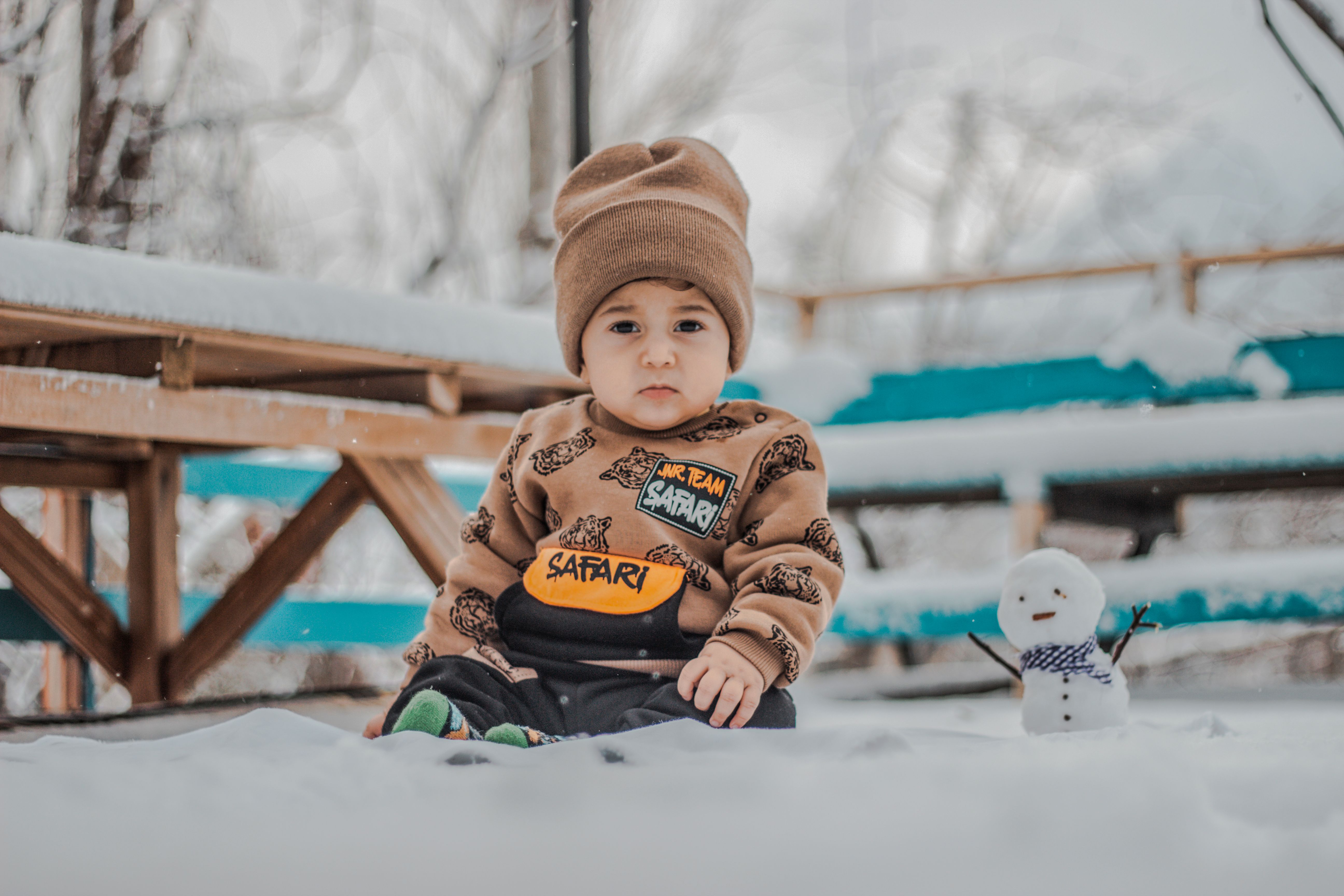 Cute child wearing winter clothes, sitting outdoors, next to a small snowman during winter.