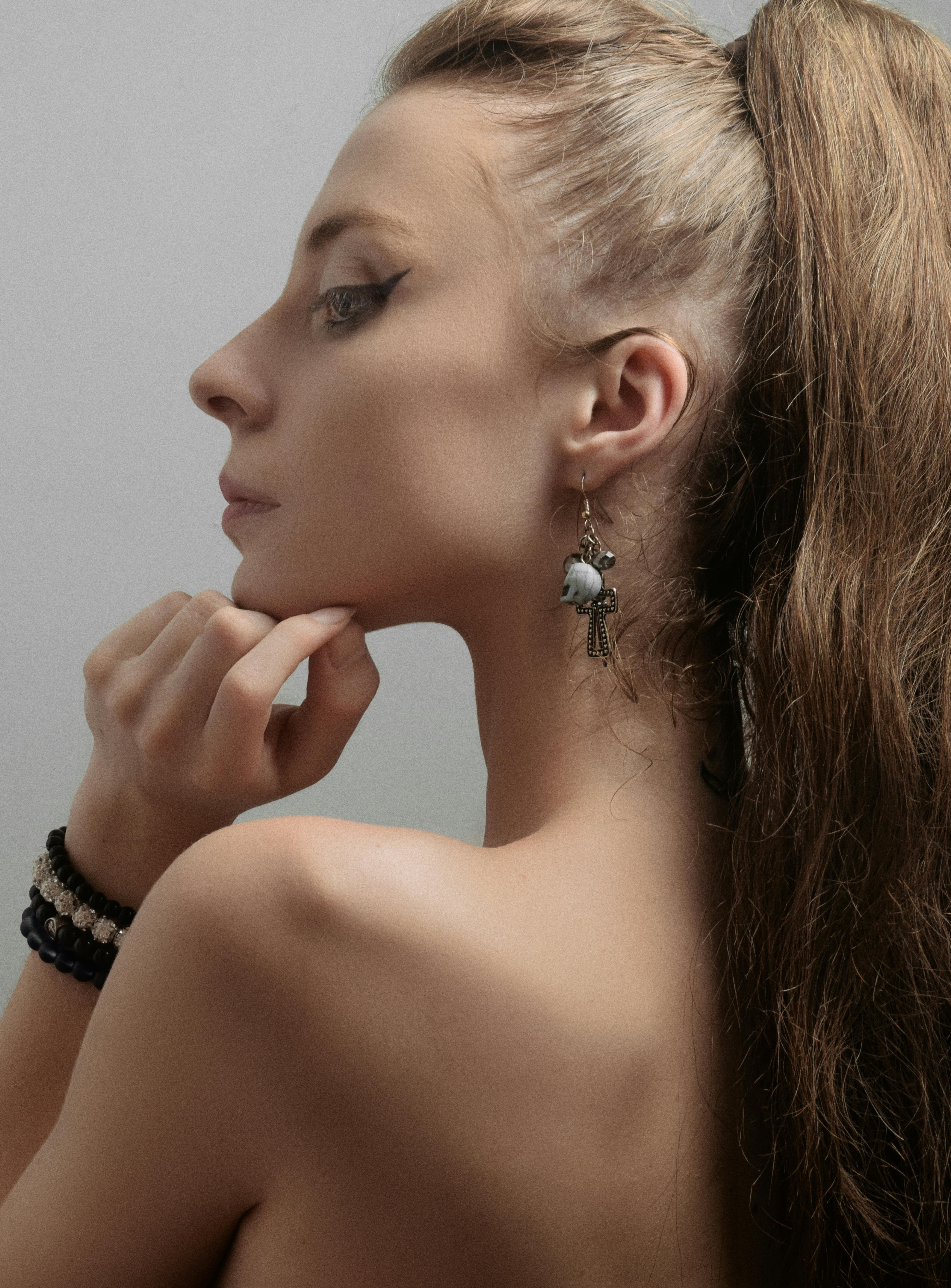 Side profile of a woman with dramatic eyeliner, showcasing earrings and a ponytail, on a grey background.