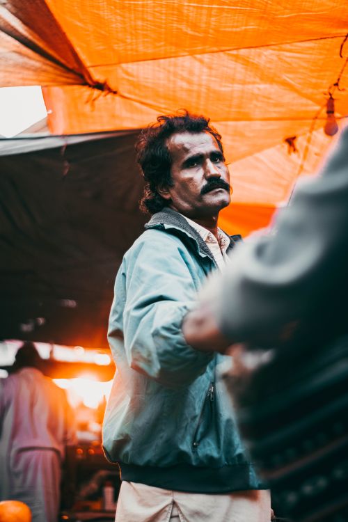 Free Portrait of a street vendor at an outdoor market under a bright orange canopy during sunset. Stock Photo