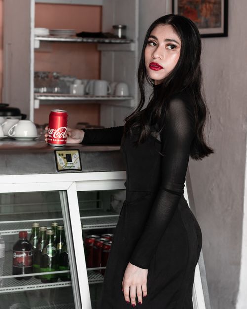 Free Stylish woman in black dress holding a soda can, posing by a café fridge for a modern lifestyle photo. Stock Photo