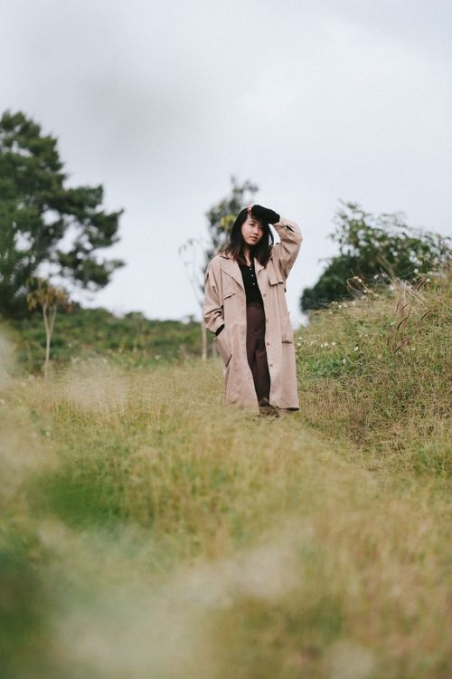 Free A stylish young woman poses in a trench coat amidst a serene countryside setting. Stock Photo
