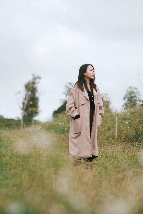 Free Woman in a beige coat standing thoughtfully in a serene rural landscape. Stock Photo