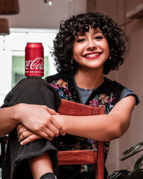 Free Happy woman with curly hair holding a Coca Cola can, sitting indoors. Stock Photo