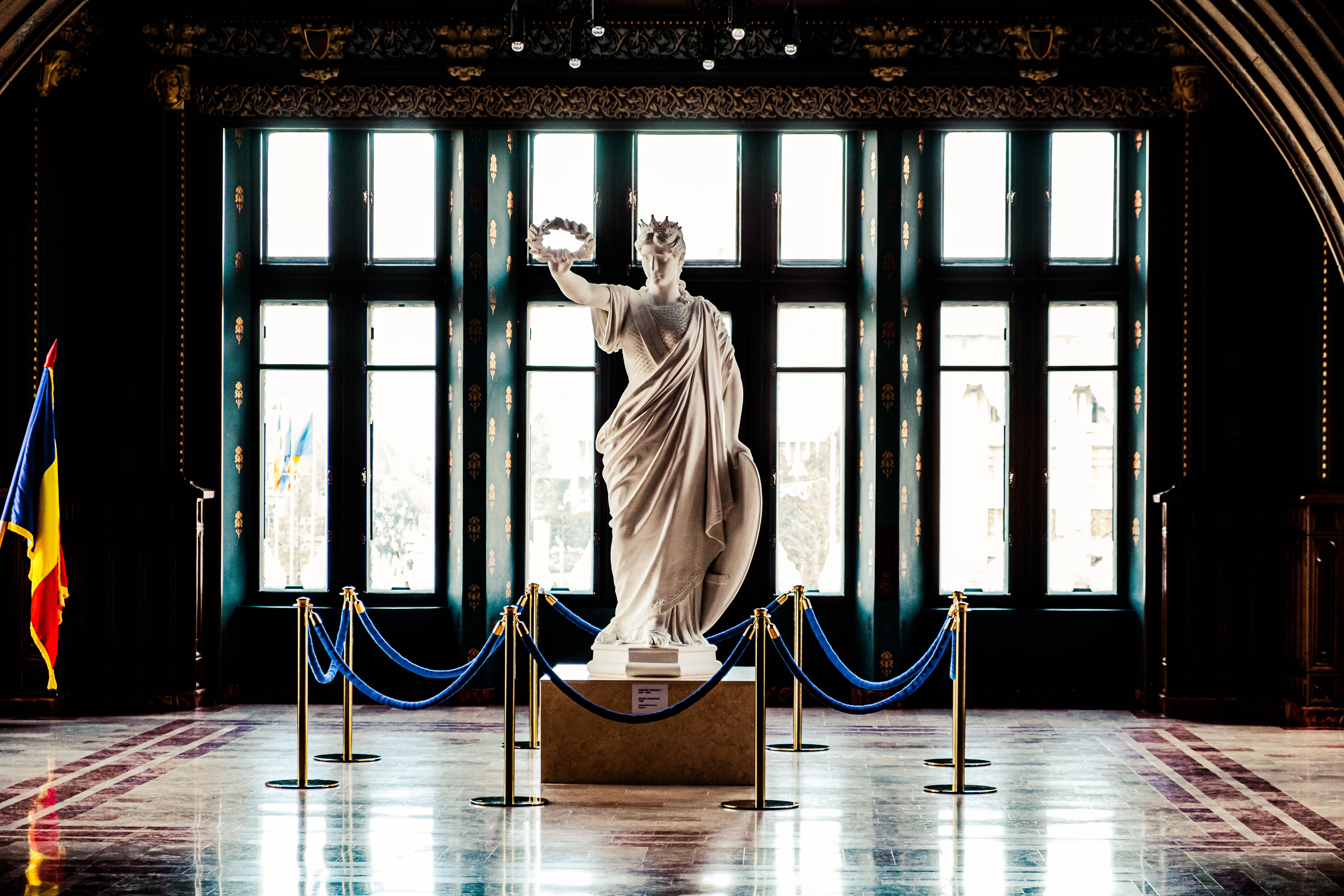 A majestic marble statue in a historic gallery with large windows and Romanian flag.