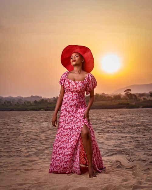 Free A stylish woman poses on a sandy beach wearing a floral dress and hat at sunset. Stock Photo