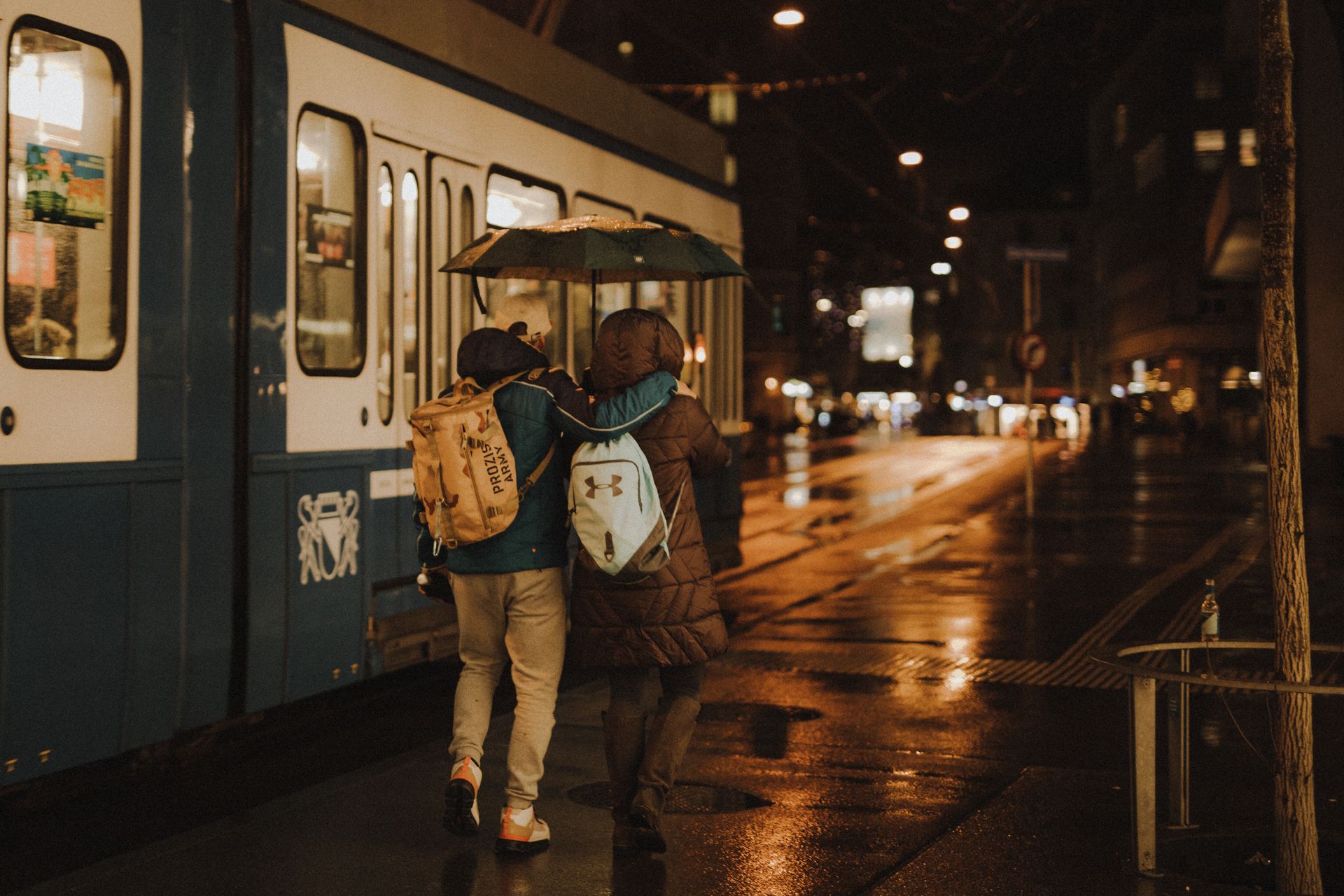 A couple walks arm in arm with umbrellas near a tram in rainy Zürich at night.