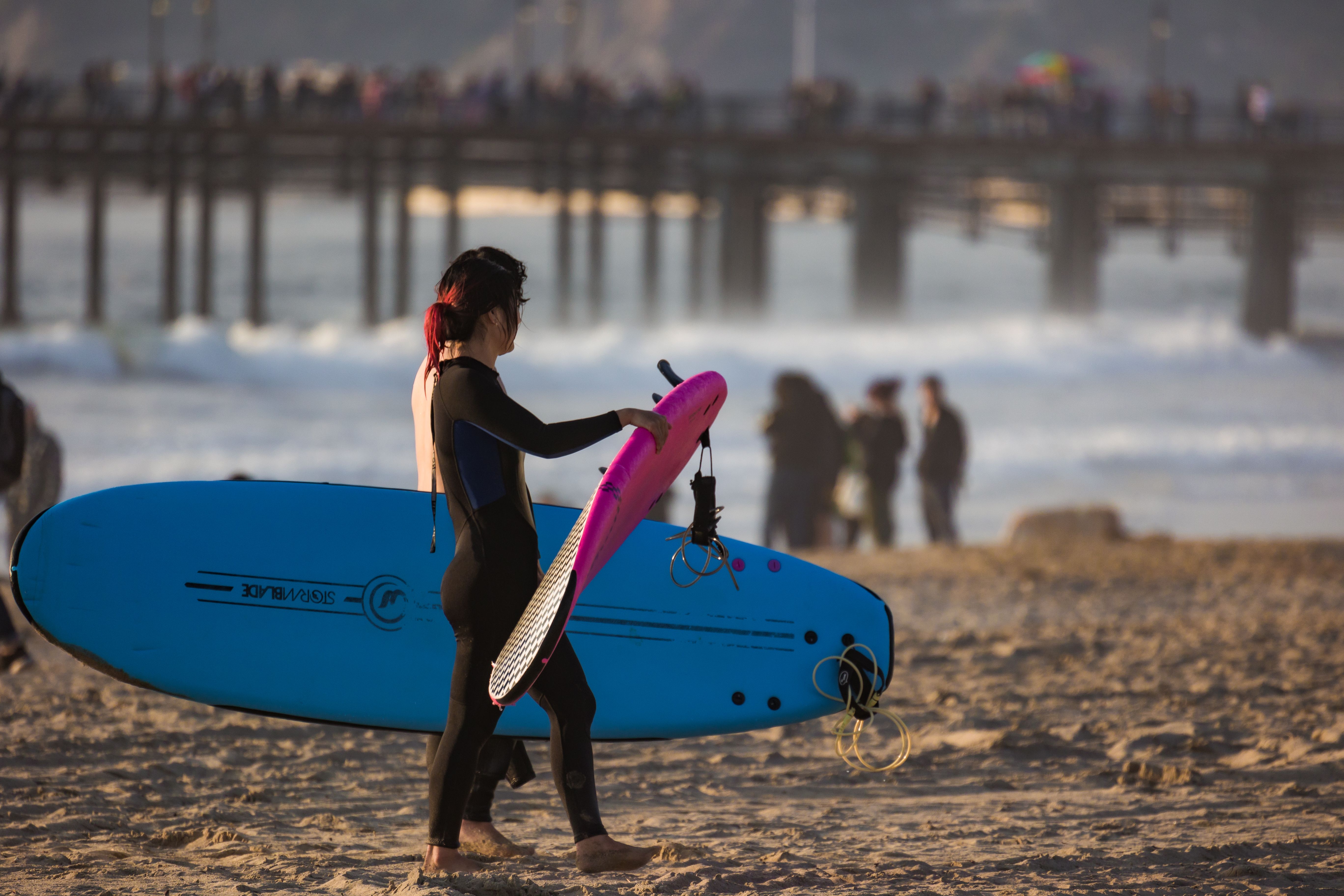 Woman carrying surfboards along a sunny beach with a pier in the background.