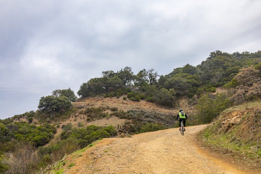 A cyclist rides along a scenic dirt road in a mountainous, rural area, offering leisure and adventure.