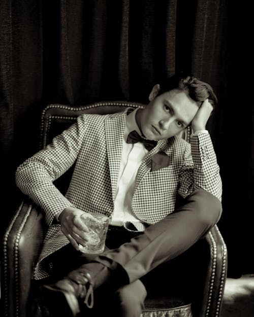 A Grayscale Photo of a Man Sitting on the Chair while Holding a Drinking Glass