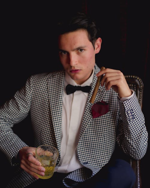 A Man in Checkered Suit Holding a Drinking Glass and Cigar while Looking with a Serious Face