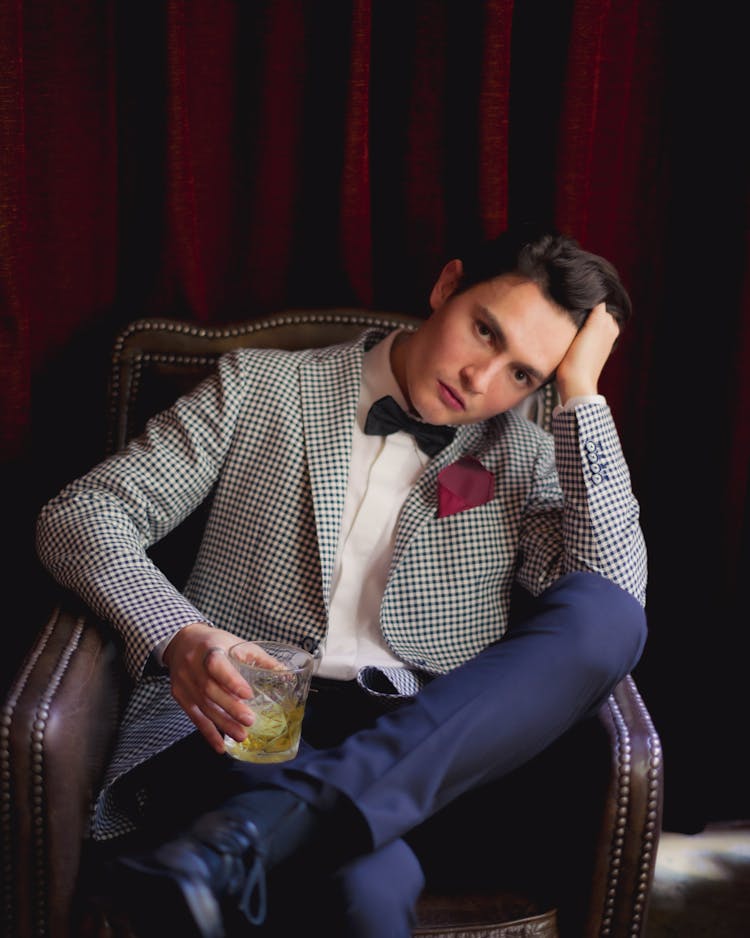 A Man In Checkered Suit Sitting On The Chair While Holding A Drinking Glass