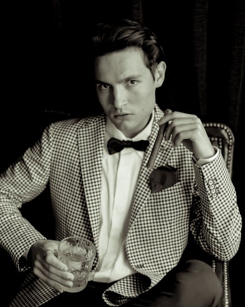 A Grayscale Photo of a Man in Checkered Suit Holding a Cigar and Whisky