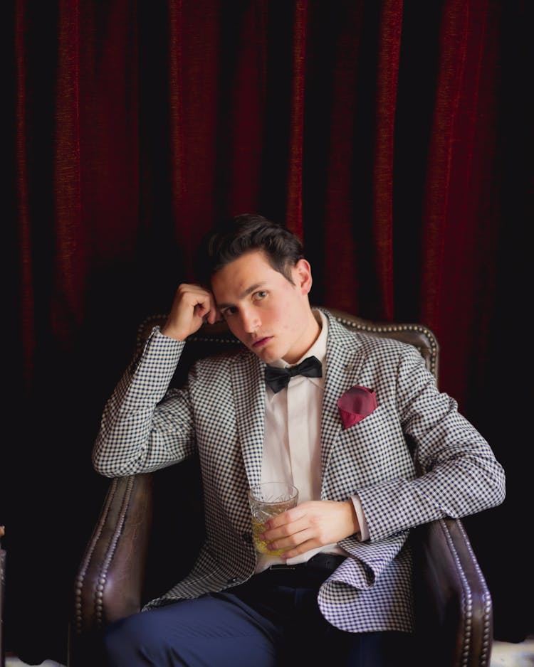 A Man In Checkered Suit Sitting On The Chair While Holding A Glass Of Whisky