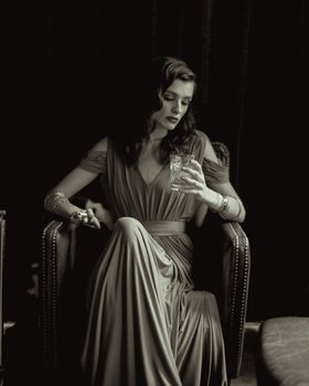 Elegant woman in a long gown holding a glass, seated in a vintage setting.