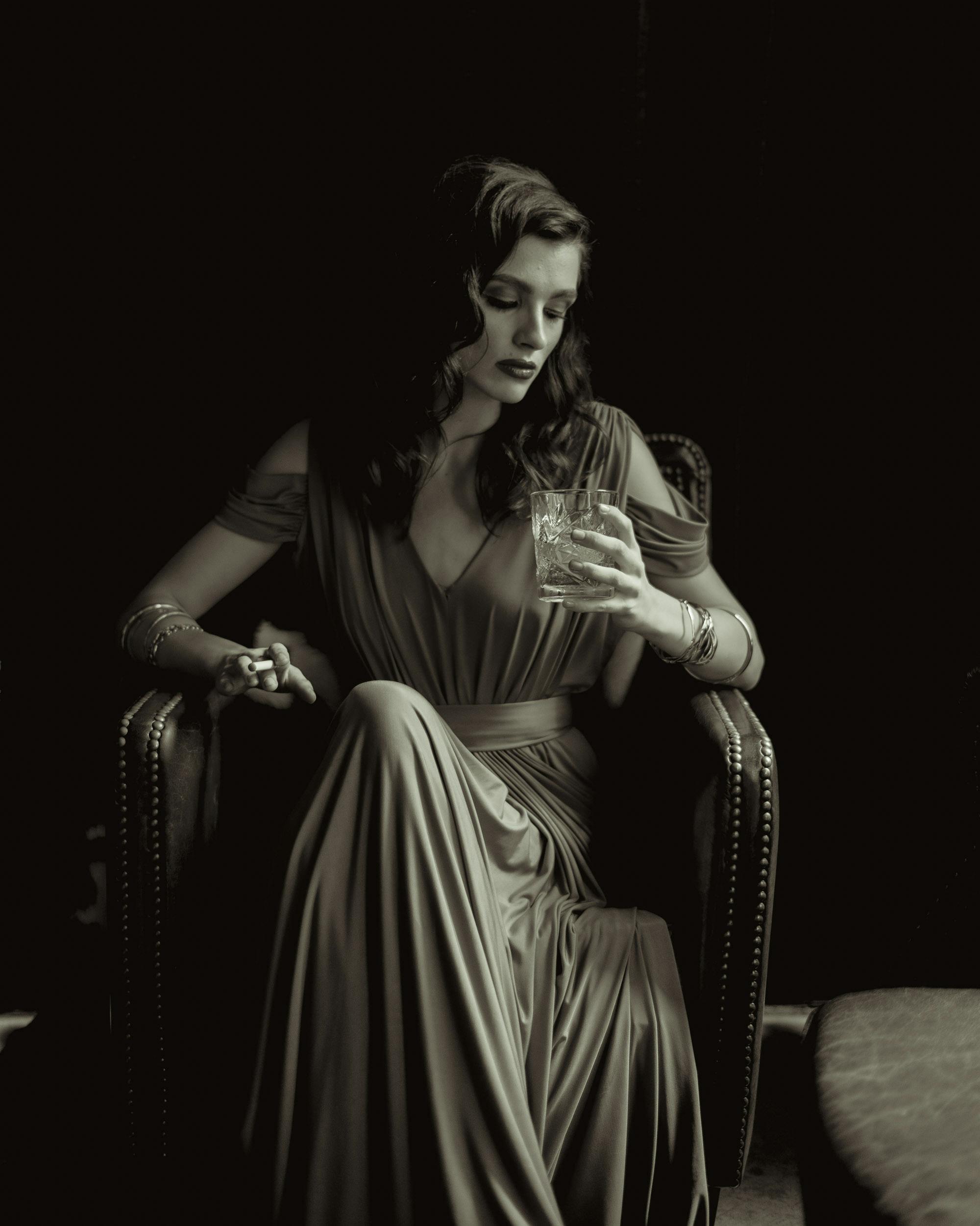 a grayscale photo of a woman sitting on the chair while holding a drinking glass