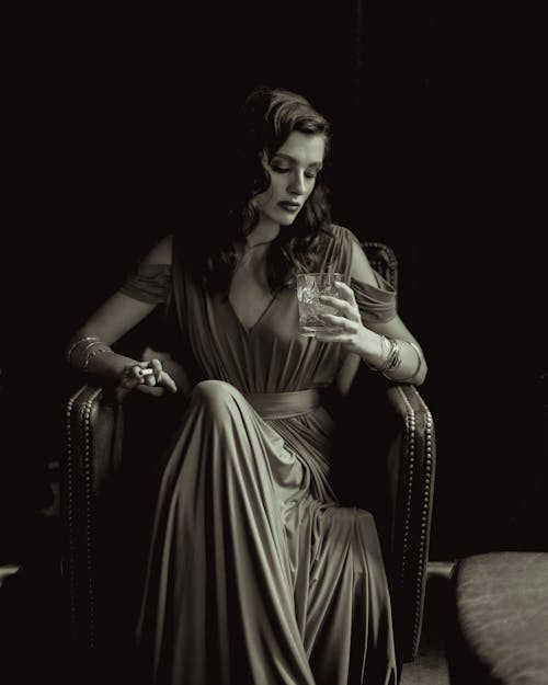 A Grayscale Photo of a Woman Sitting on the Chair while Holding a Drinking Glass