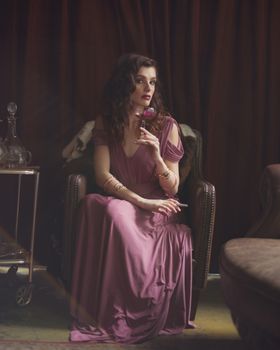 A woman in a purple dress sits elegantly in a dimly lit room, holding a glass of wine.