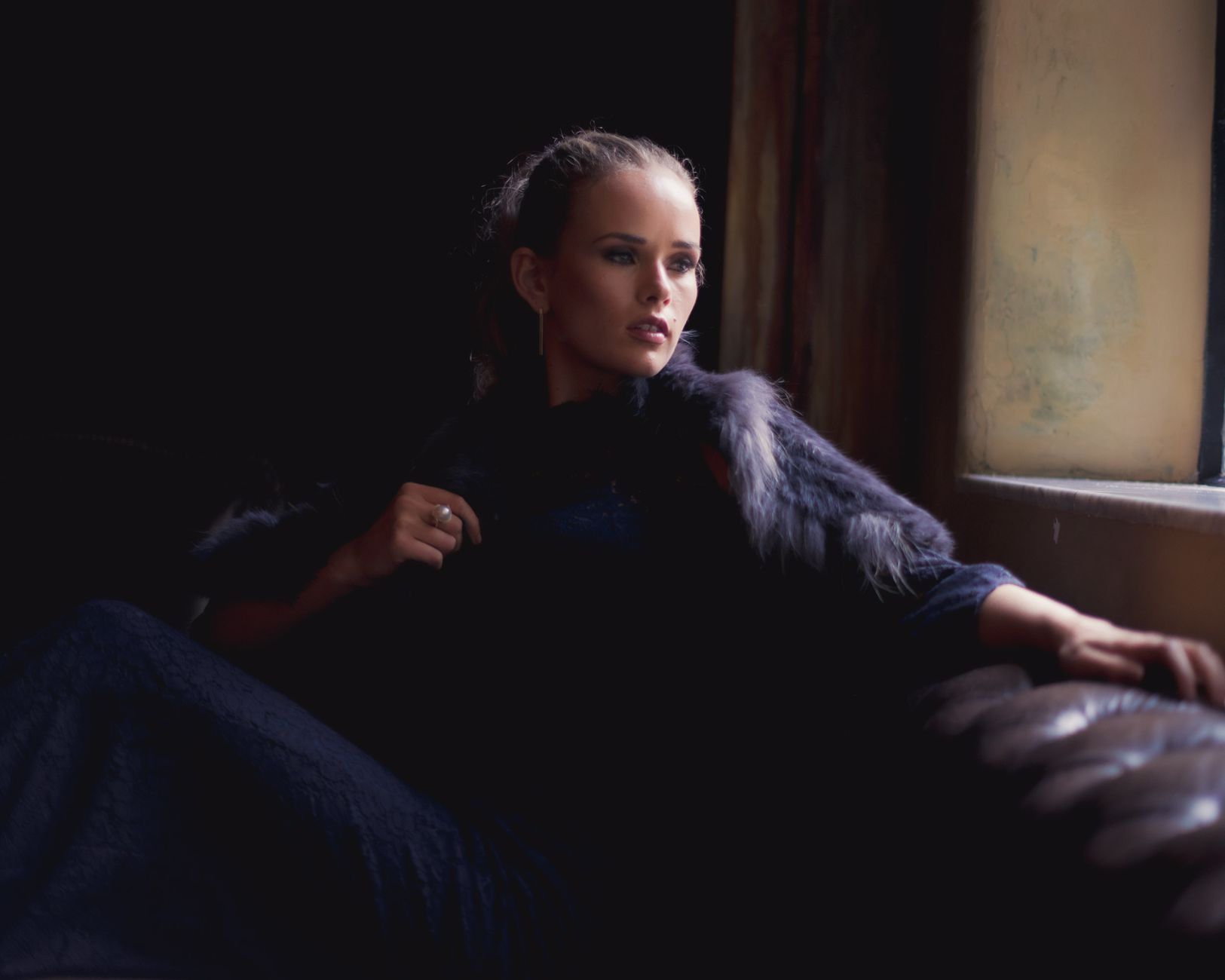 Portrait of a stylish woman in fur coat relaxing by a window in a dim luxury interior.