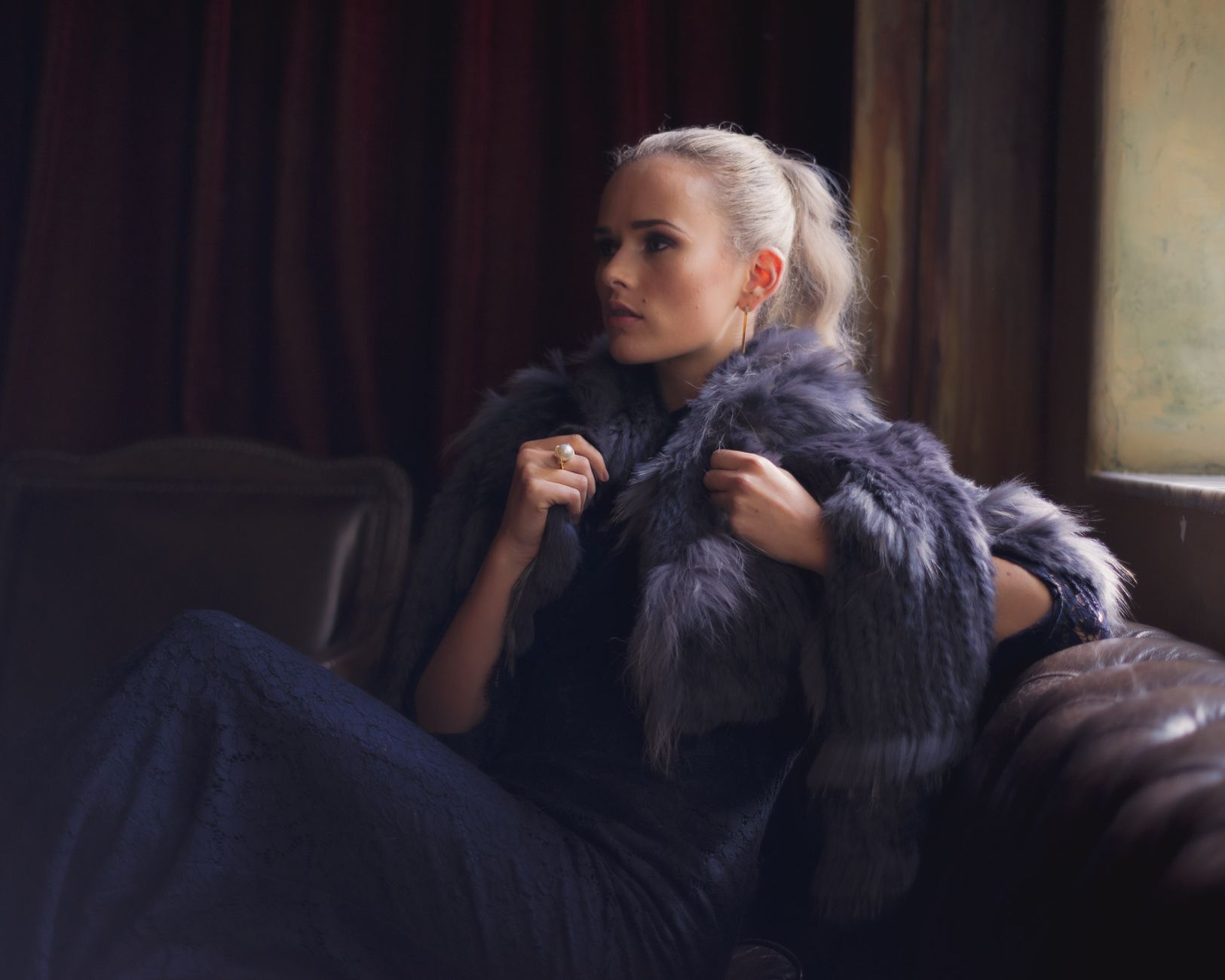 Elegant woman in a blue lace dress with a fur coat in a luxurious interior setting.