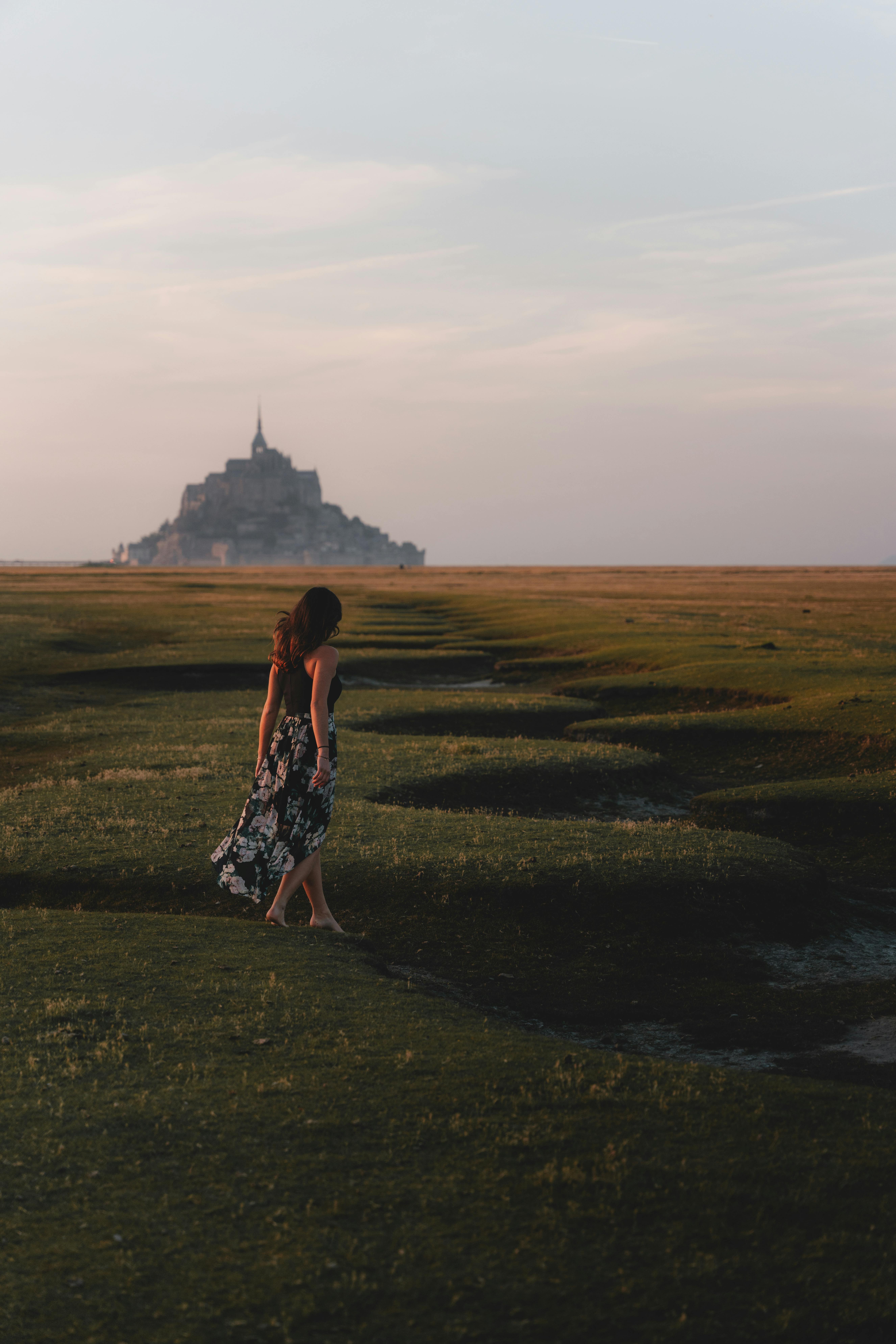 A woman in a floral dress walks towards Le Mont-Saint-Michel at sunset in the French countryside.