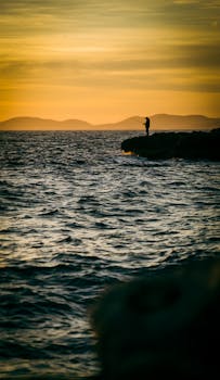 A lone angler silhouetted against a vibrant sunset on a rocky seashore, casting into the ocean.
