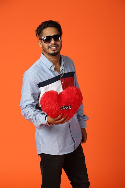 Fashionable man posing with a red heart pillow against an orange backdrop, embodying joy and love.